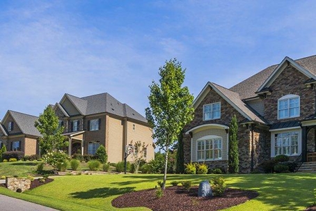 Two nice large homes with a wide green yard with shrubs and small trees stretched in front of them.